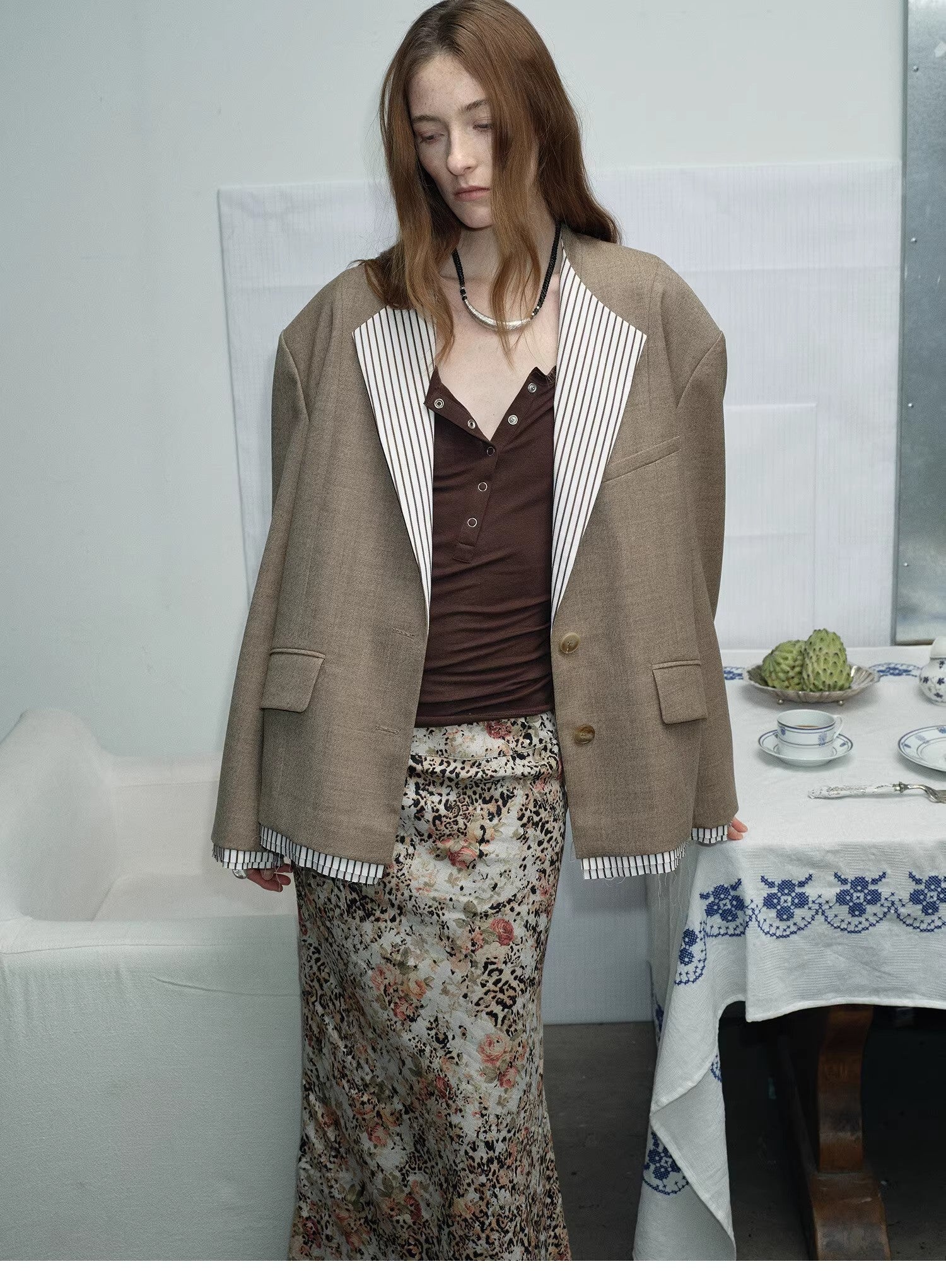 Woman wearing a brown blazer layered over a floral skirt, standing in a decorated room near a table. This smart-casual lesbian outfit blends professional lesbian style with authentic lesbian fashion.
