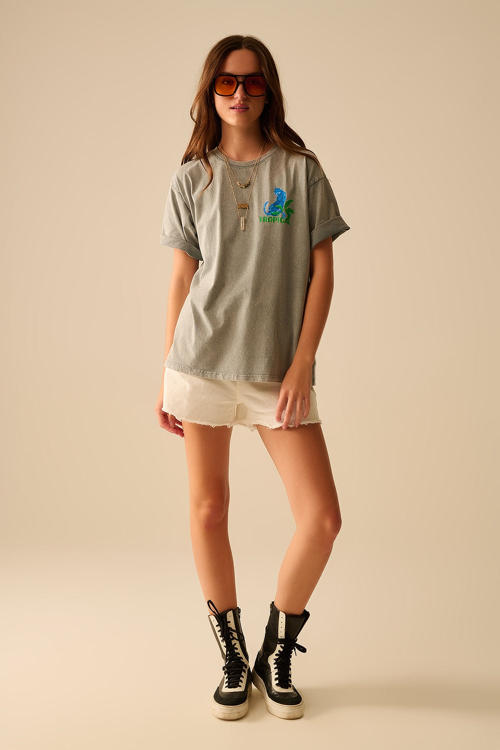 Woman wearing a gray t-shirt with a graphic design, white shorts, and sharp black boots on a beige background. This confident lesbian outfit is a perfect example of casual lesbian fashion and authentic lesbian style.