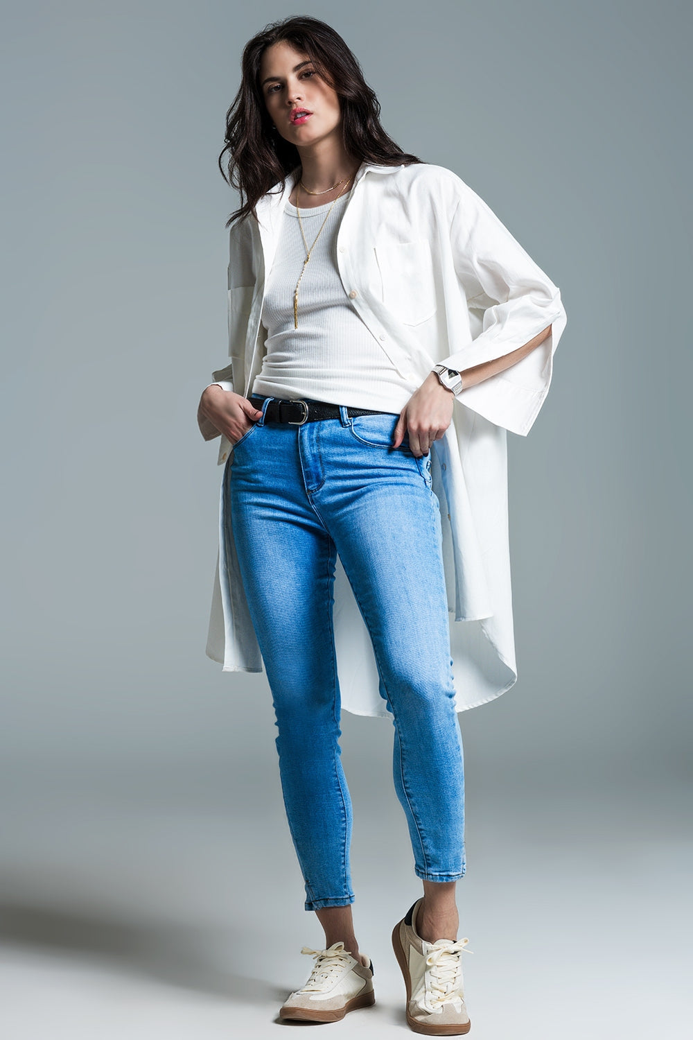 Woman wearing a white shirt, blue jeans, and a long white coat on a gray background. This sharp, layered lesbian outfit is a perfect example of confident lesbian fashion and chic lesbian style.