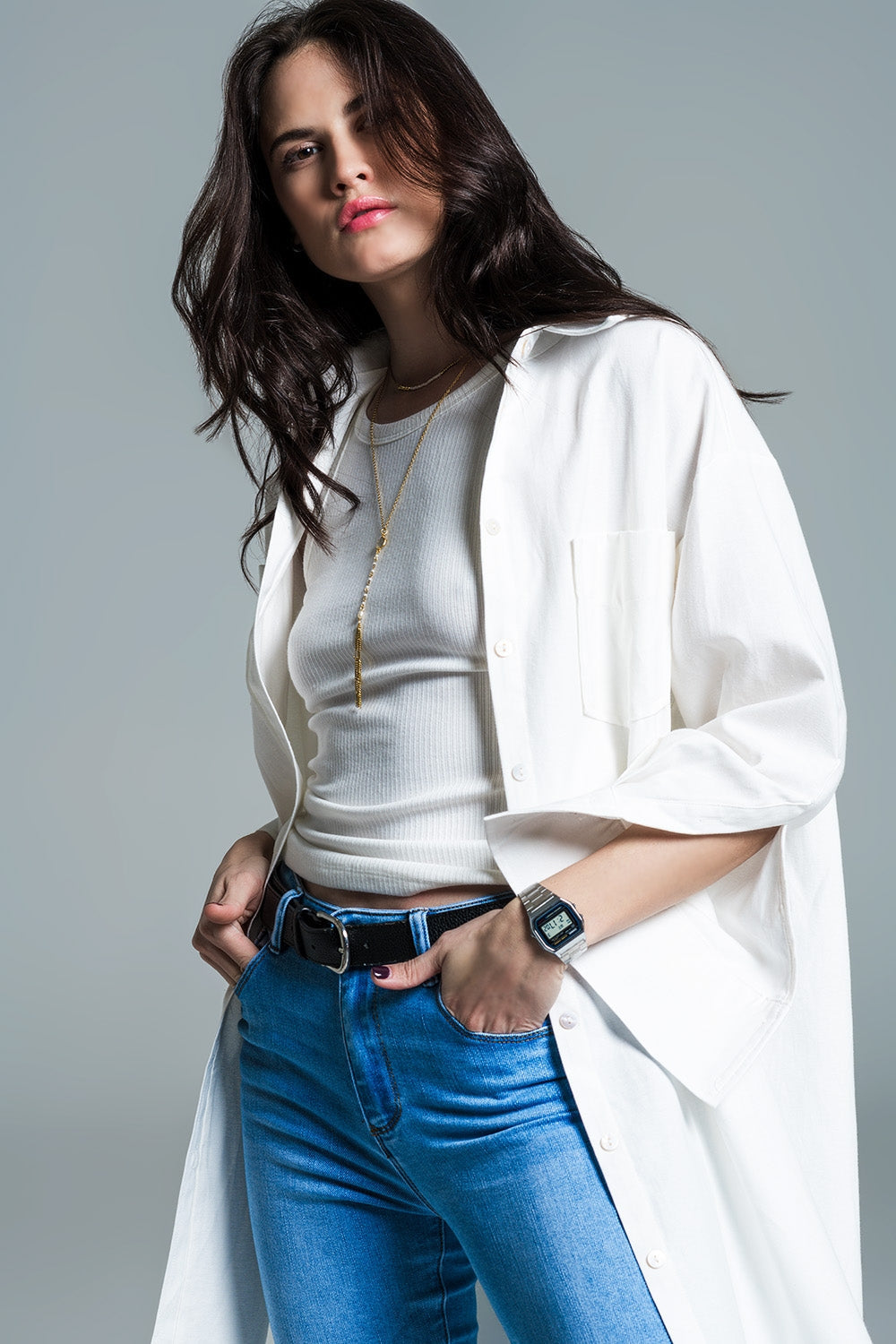 Woman wearing a long white coat layered over a white shirt and blue jeans on a gray background. This sharp, minimalist lesbian outfit is a perfect example of confident lesbian fashion and chic lesbian style.