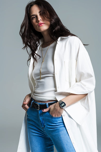 Woman wearing a long white coat layered over a white shirt and blue jeans on a gray background. This sharp, minimalist lesbian outfit is a perfect example of confident lesbian fashion and chic lesbian style.