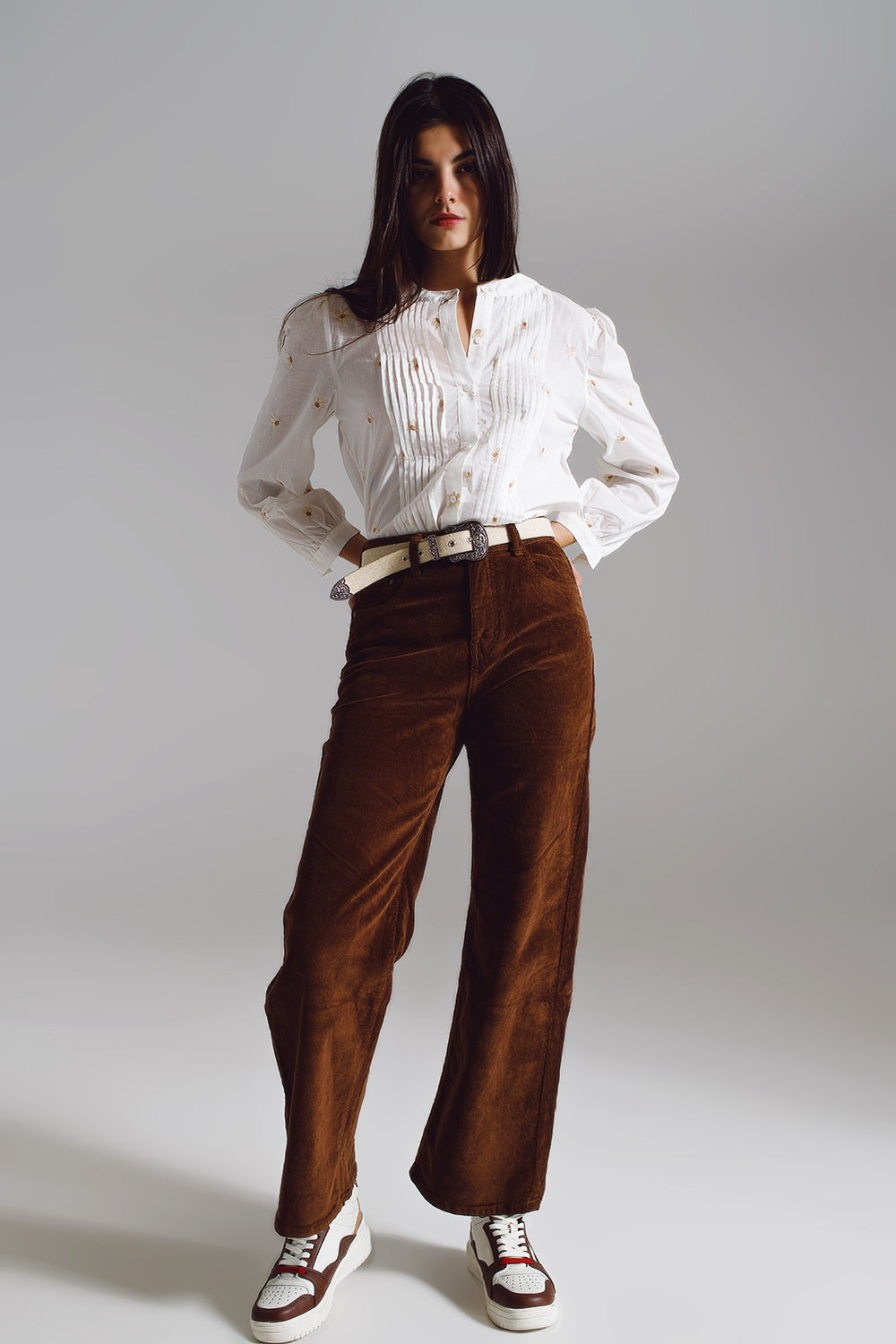 Woman modeling a simple, professional lesbian outfit featuring a white blouse and brown pants on a plain background. This look is a strong example of everyday lesbian fashion and clean lesbian style.